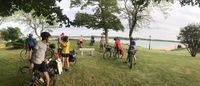A group of cyclists on touring bikes, gathered in a park in front of a canal. Nearly all of them are facing the camera, but looking back at a rider in the far middle of the shot.