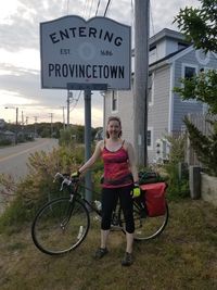 Shannon, in cycling gear, posing with her bike by the "Entering Provincetown" sign on Route 6A.